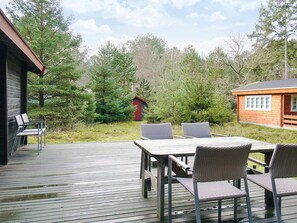 Una terraza de madera con una mesa y sillas, rodeada de árboles y un pequeño cobertizo rojo.
