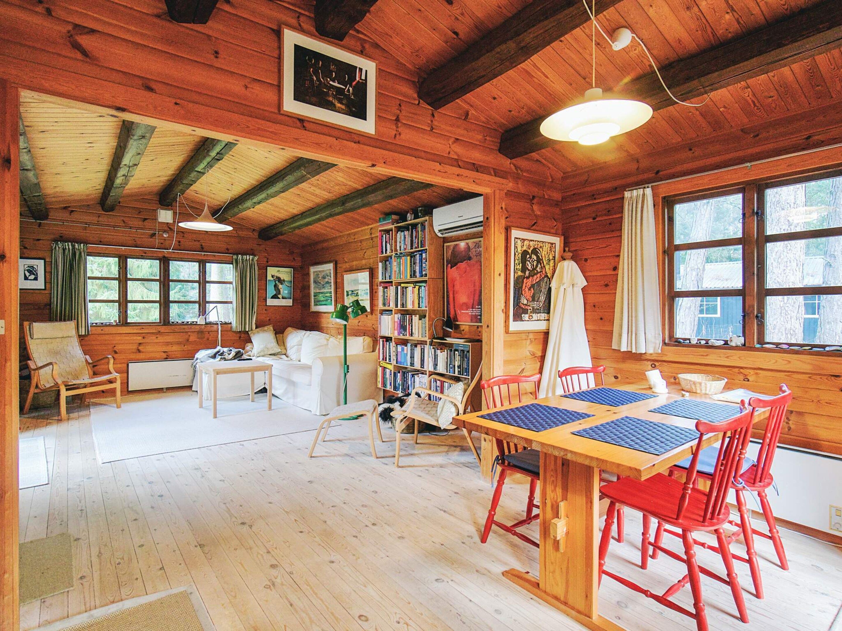 A cozy cabin with a wooden interior, a dining area with red chairs, a bookshelf, and large windows.