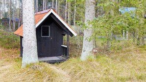 Une petite cabane en bois sombre, avec un toit en pente, entourée de grands arbres et d’une prairie.