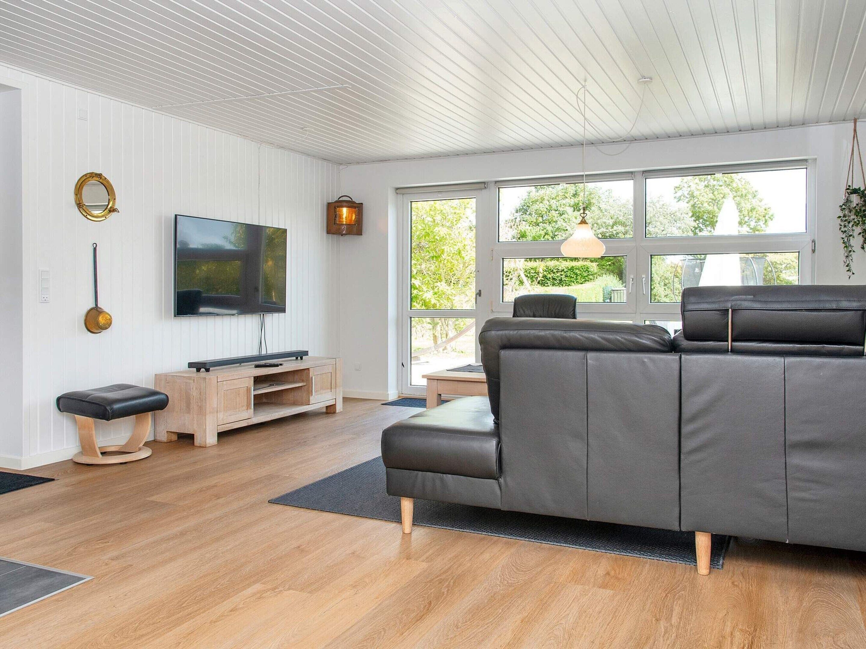 A modern living room with a large grey sofa, a flat-screen TV, wooden flooring, and large windows offering a view of greenery outside.