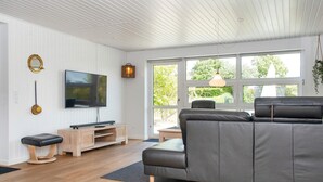 Un salon moderne doté d’un grand canapé gris, d’un téléviseur à écran plat, d’un parquet et de grandes fenêtres offrant une vue sur la verdure environnante.