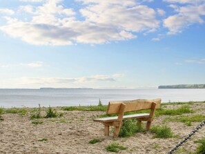 A wooden bench on a sandy beach with a view of the sea and a distant landmass. - 7 Person Holiday Home in Bjert (Bjert)