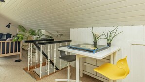 Una habitación con techo blanco, pared blanca, puerta blanca, una mesa blanca con un libro y dos plantas en macetas, una silla amarilla, una silla negra, una lámpara de pie negra y una escalera con balaustres blancos y barandilla negra.