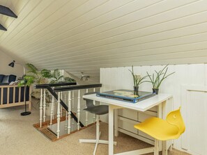 A room with a white ceiling, a white wall, a white door, a white table with a book and two potted plants, a yellow chair, a black chair, a black floor lamp, a staircase with white balusters, and a black railing.