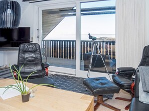 A room with a sliding glass door leading to a balcony, a wooden table, two leather chairs, a tripod, and a television.