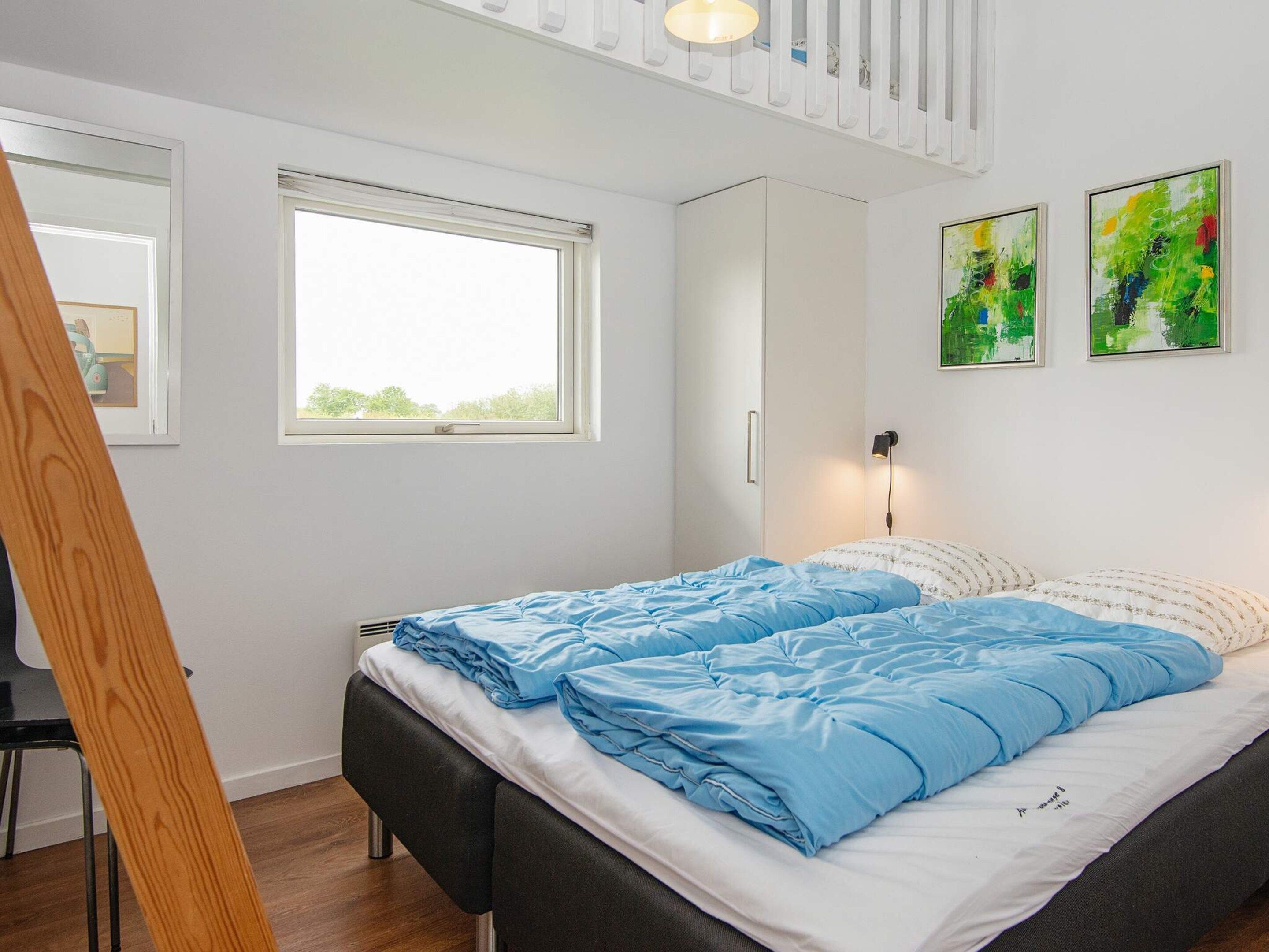 A bedroom with a bed, a window, a wall-mounted light, and two abstract paintings.