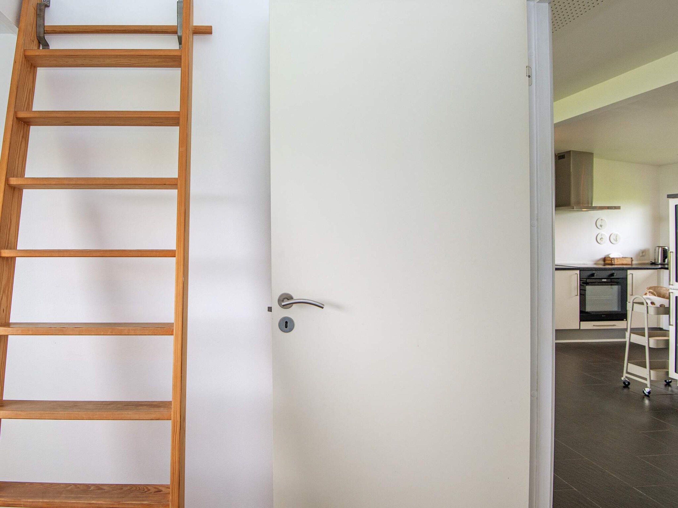 A white door with a silver handle, a wooden ladder leaning against it, and a glimpse of a kitchen area with cabinets and appliances.