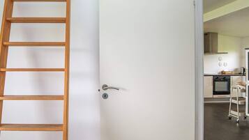 A white door with a silver handle, a wooden ladder leaning against it, and a glimpse of a kitchen area with cabinets and appliances.