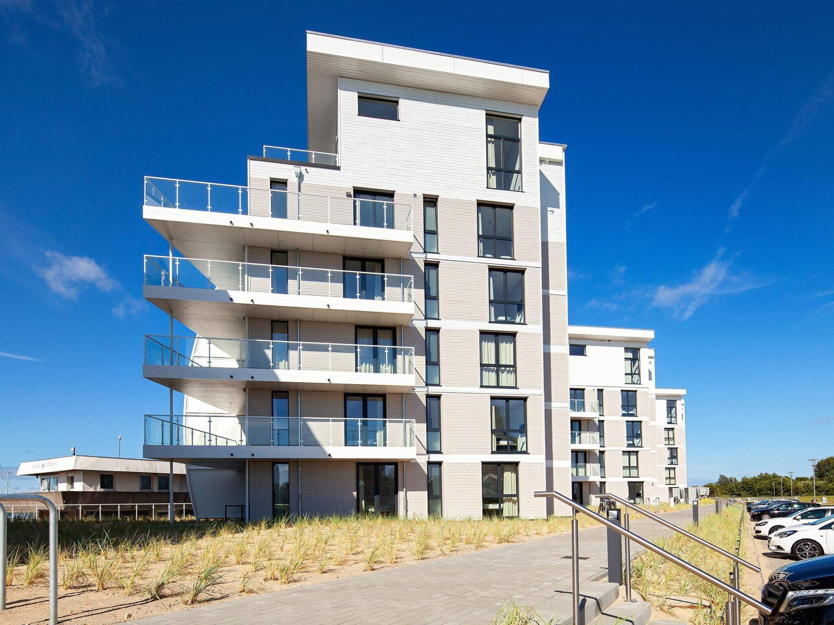 A multi-story residential building with balconies, a parking area, and a clear sky.