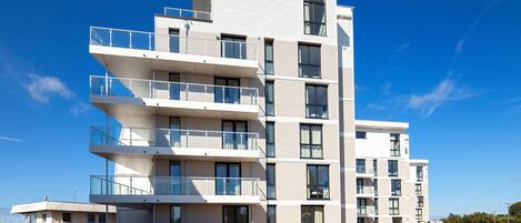 A multi-story residential building with balconies, a parking area, and a clear sky.
