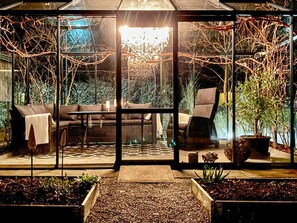 A glass-enclosed outdoor seating area with a sofa, chairs, and a table, surrounded by plants and a decorative tree.