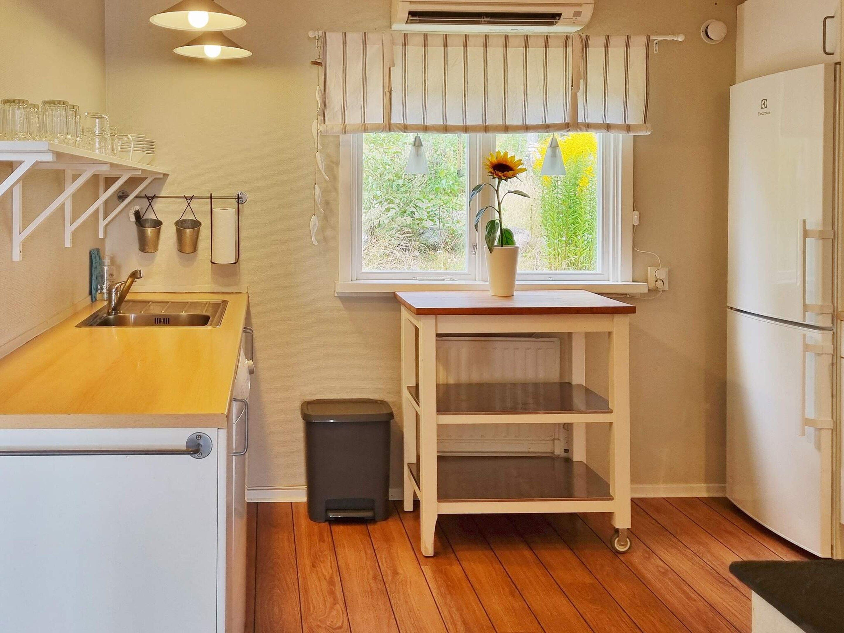 A kitchen with a wooden countertop, a sink, a trash bin, a window with a sunflower, and a refrigerator.