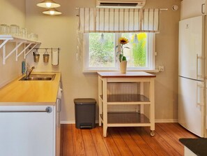 Una cocina con encimera de madera, un lavamanos, un cubo de basura, una ventana con un girasol y un refrigerador.