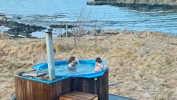 A hot tub with two people in it, surrounded by a grassy area and a body of water with mountains in the background. - 8 Person Holiday Home in Bolandet-by Traum (Heroy)