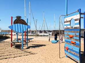 A beach with a climbing structure, a shaded area, and boats in the background. - 5 Star Holiday Home in Wendtorf (Wendtorf)