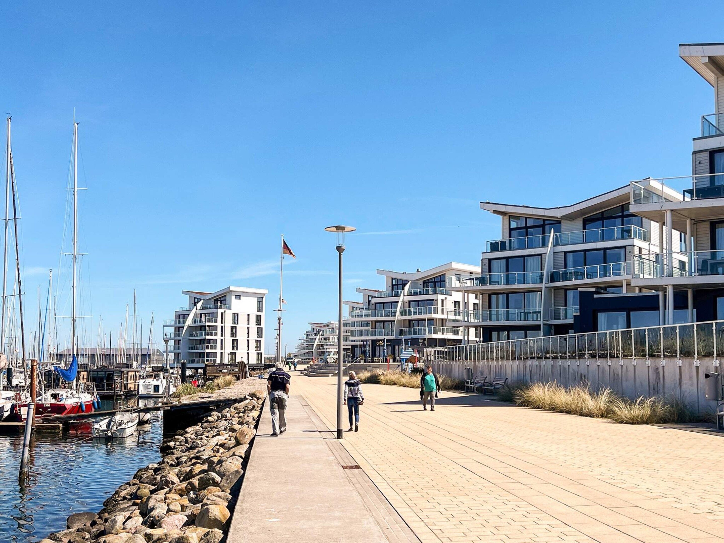 A waterfront promenade with modern buildings, a marina with sailboats, and a clear sky.