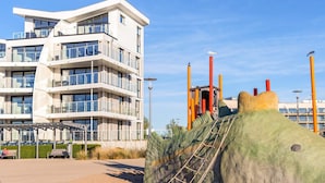 A modern multi-story building with balconies, a playground with climbing structures, and a sandy area with benches. - 5 Star Holiday Home in Wendtorf (Wendtorf)