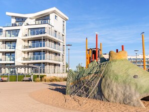 En modern, flervåningsbyggnad med balkonger, en lekplats med klätterställningar och ett sandområde med bänkar.