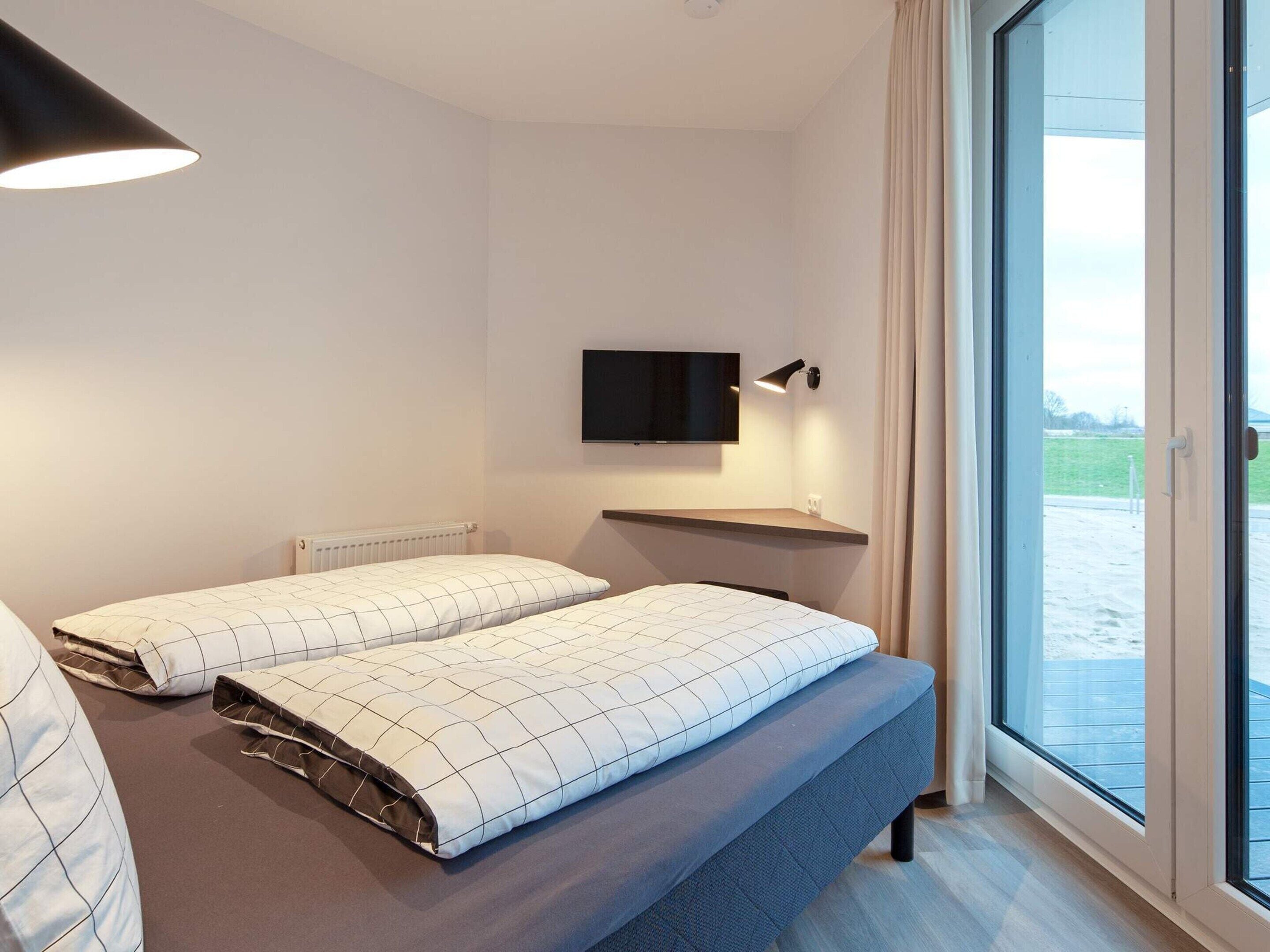 A modern hotel room with a bed, a TV mounted on the wall, and a large window offering a view of the outdoors.