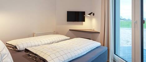A modern hotel room with a bed, a TV mounted on the wall, and a large window offering a view of the outdoors.