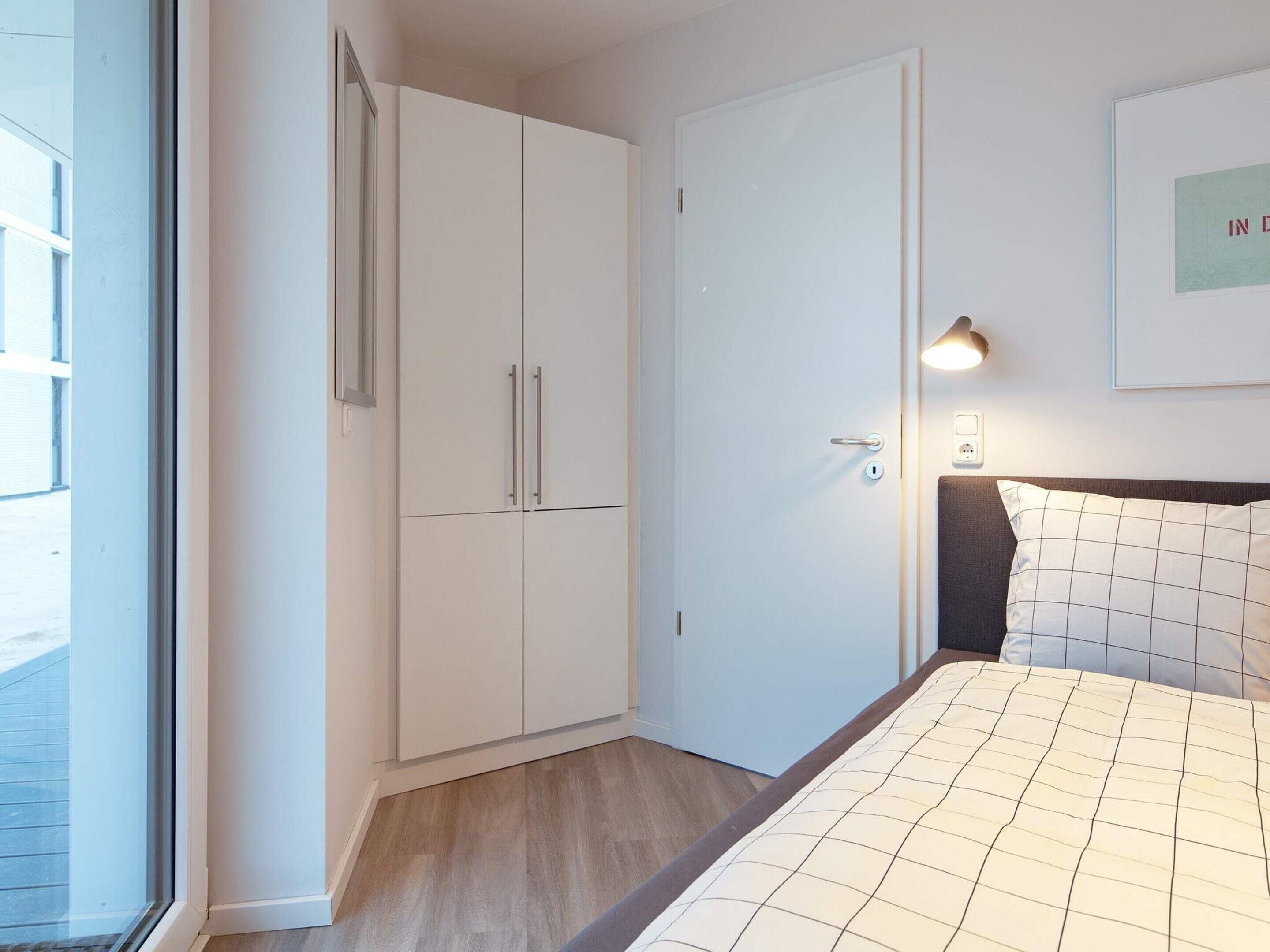 Quarto com cama, guarda-roupa, porta, luminária na parede e quadro emoldurado.