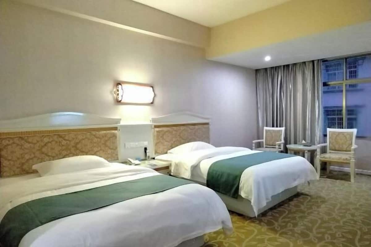 Quarto de hotel com duas camas, mesa de cabeceira, uma cadeira, abajur e janela com cortinas.