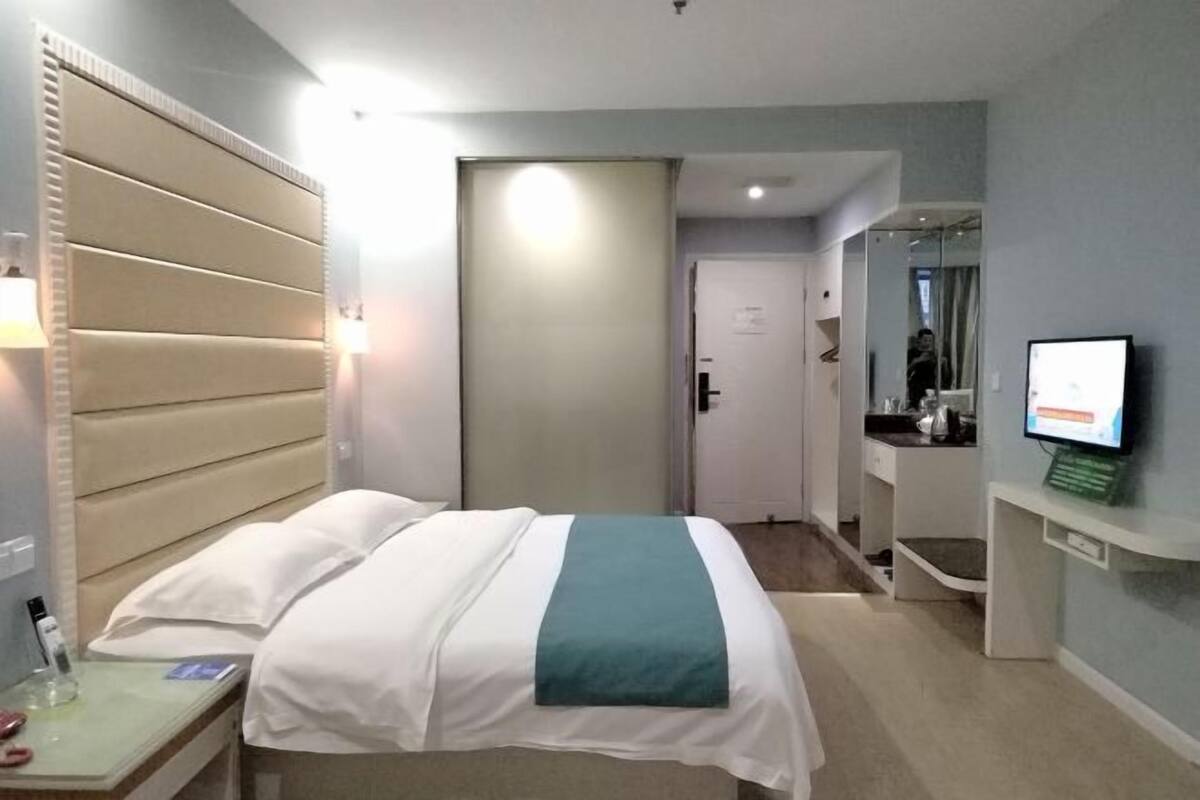 Quarto de hotel com cama, mesa de cabeceira, luminária, televisão e porta para o banheiro.