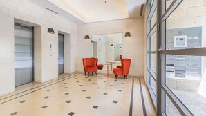 Un hall d’entrée avec un sol carrelé, deux ascenseurs, des chaises rouges, un mur en verre offrant une vue et un miroir.