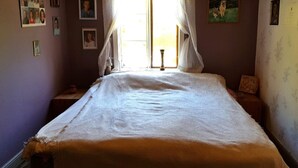 A bedroom with a large bed, a window with lace curtains, and various framed pictures on the walls.