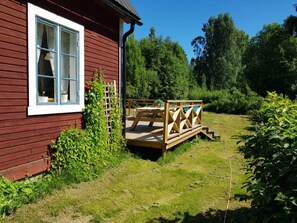 A red wooden cabin with a white window, a wooden deck, and a garden area.