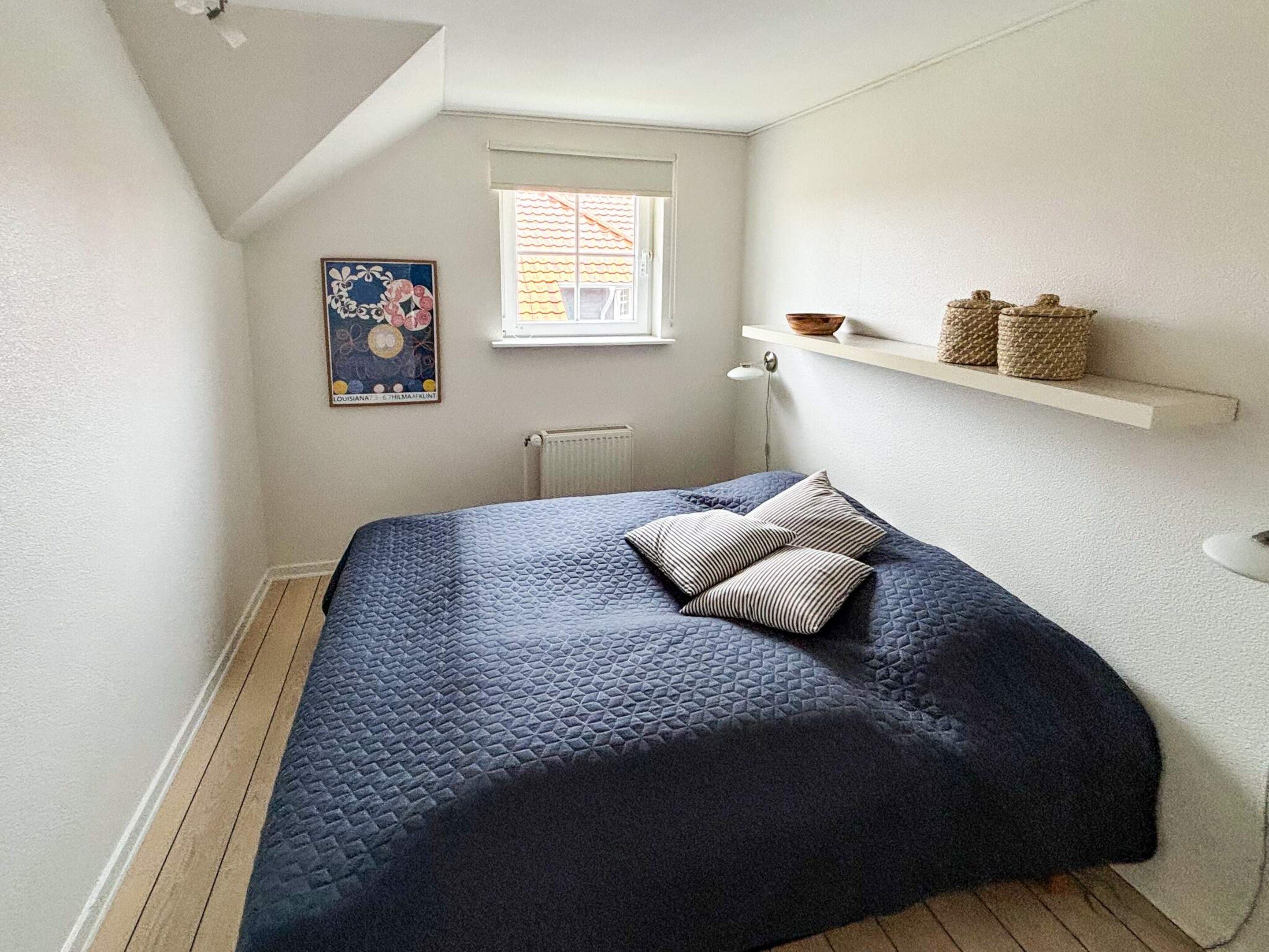 Un dormitorio con cama, ventana, radiador y una estantería con adornos.