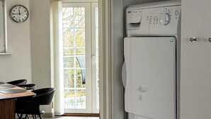 A room with a large window, a wall clock, a washing machine, and a dining area with chairs and a table. - 4 Star Holiday Home in Bogense (Bogense)