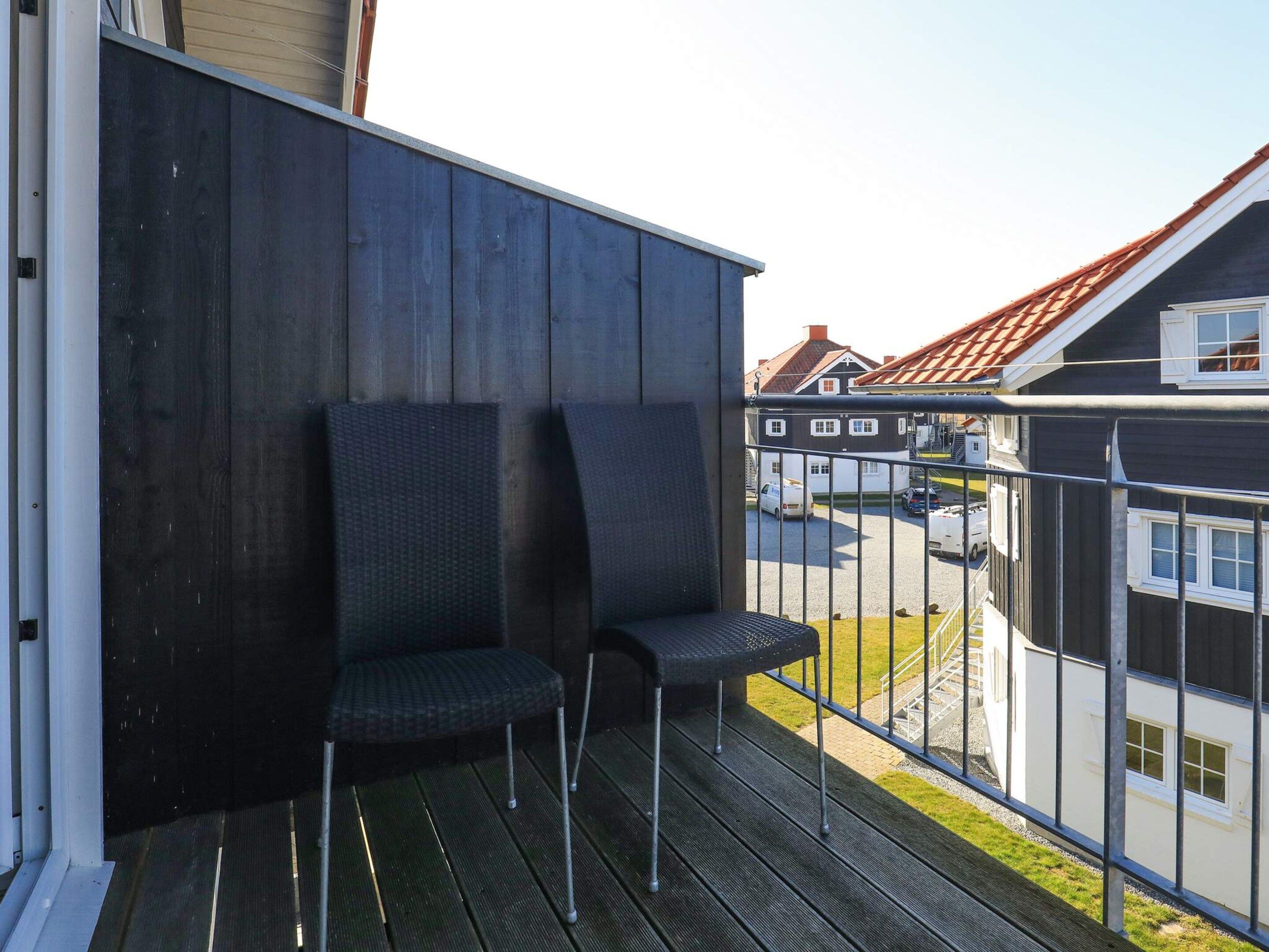 Ein Balkon mit zwei Stühlen und einem Metallgeländer, der auf ein Wohngebiet mit Häusern und einem klaren Himmel blickt.