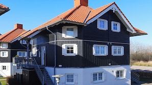 Ein mehrstöckiges Gebäude mit dunkler Fassade, weißem Zierrat und roten Ziegeldächern. Es hat mehrere Fenster, eine sichtbare Treppe und eine gepflasterte Fläche davor.