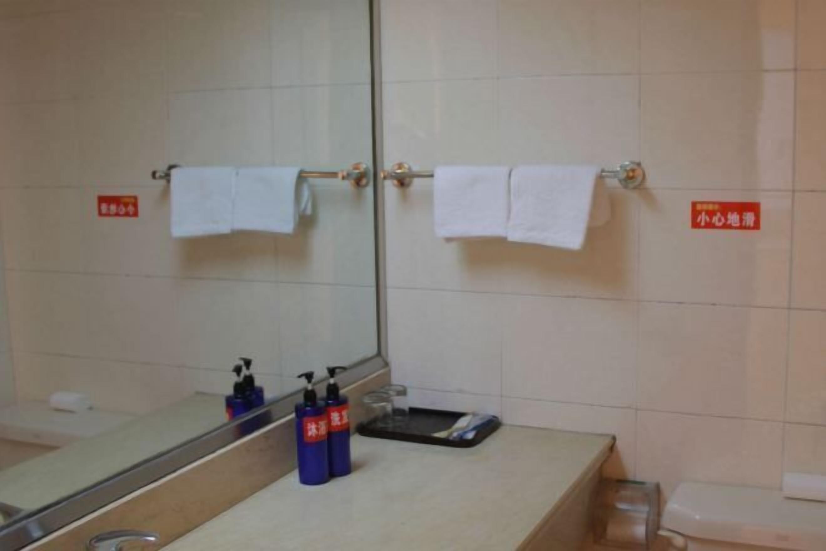 A bathroom with a mirror, two towels hanging on a towel rack, a sink with soap dispensers, and a trash bin.