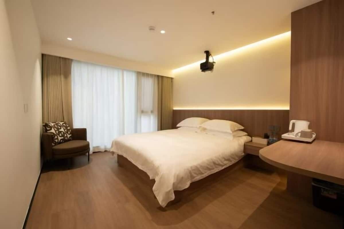A hotel room with a large bed, a bedside table, a chair, and a desk with a lamp.