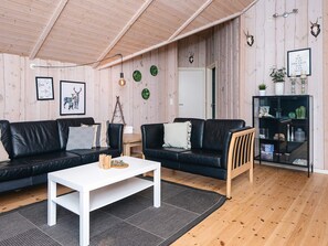 A modern living room with two black leather sofas, a white coffee table, and a wooden chair. There is a bookshelf with decorative items and a door leading to another room.