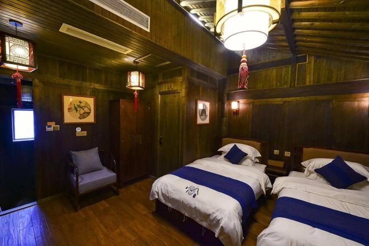 A room with wooden walls, two beds with blue bedding, a chair, and a ceiling light fixture.