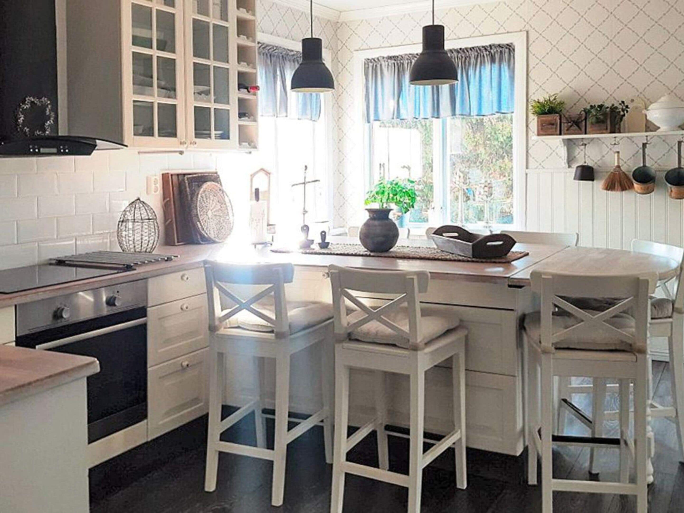 A kitchen with white cabinetry, a central island with stools, pendant lighting, and a window with curtains.