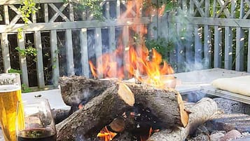 A campfire with logs and a pot, surrounded by a wooden fence and greenery.