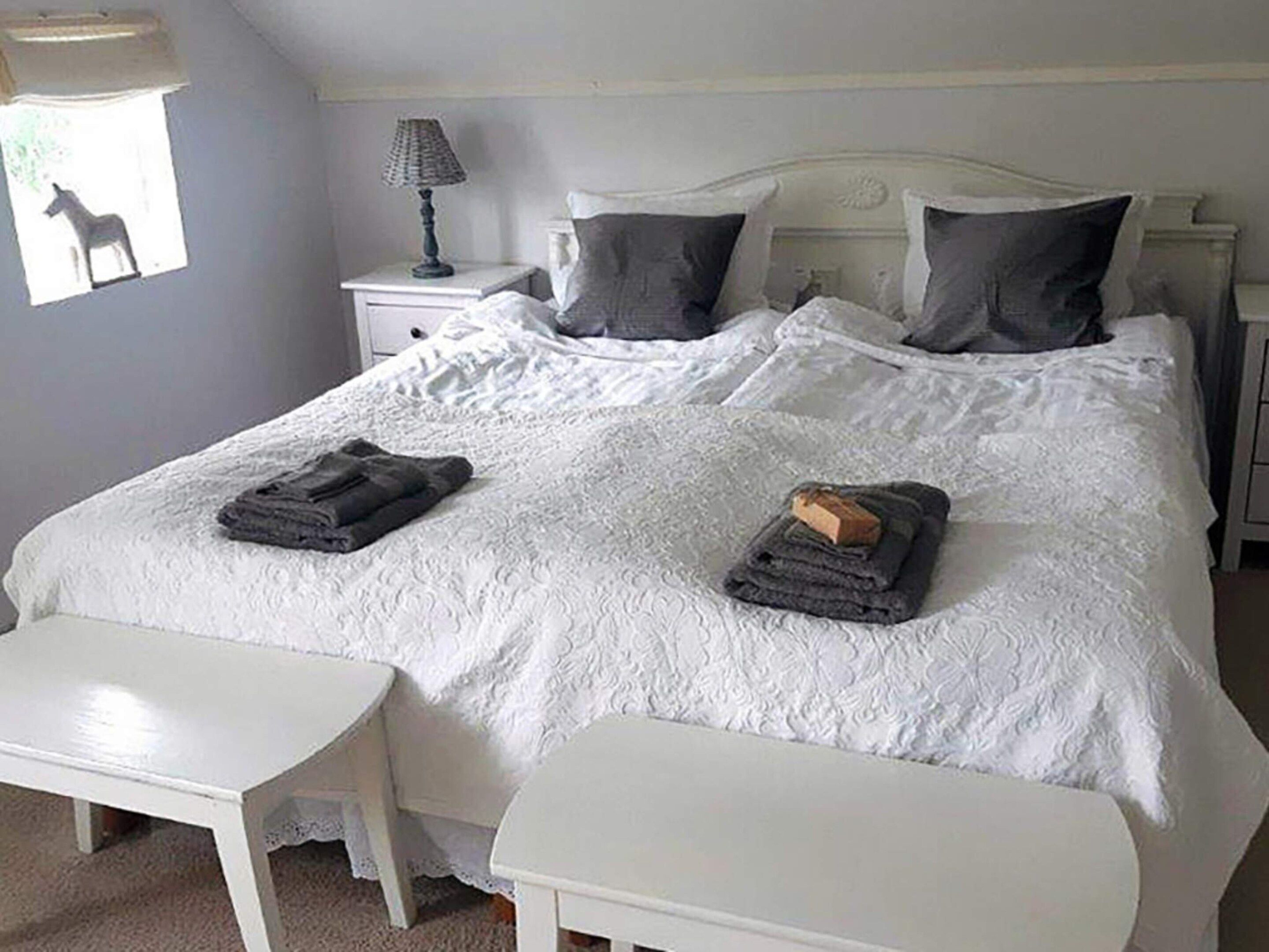 A double bed with a white duvet, two pillows, a bedside table with a lamp, and a small chair.