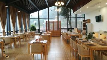A spacious dining area with multiple wooden tables and chairs, large windows, and a modern ceiling design.