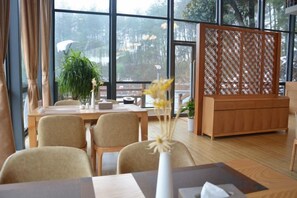 A room with a wooden dining table, chairs, a vase with flowers, and a large window with a view of trees. - Welcome Song Hotel (Huangshan City)