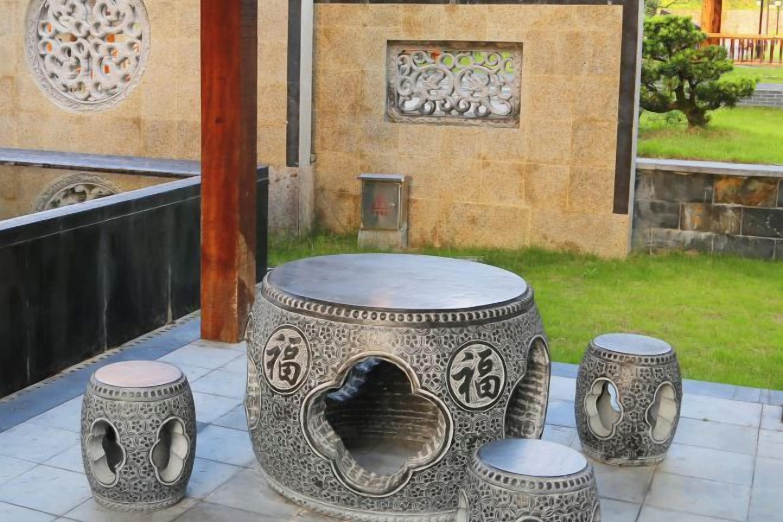 A courtyard with a stone wall, a round table with intricate carvings, and four matching stools.