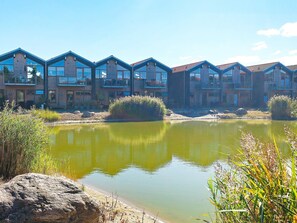 A row of modern multi-story buildings with balconies, a small pond, and surrounding vegetation.