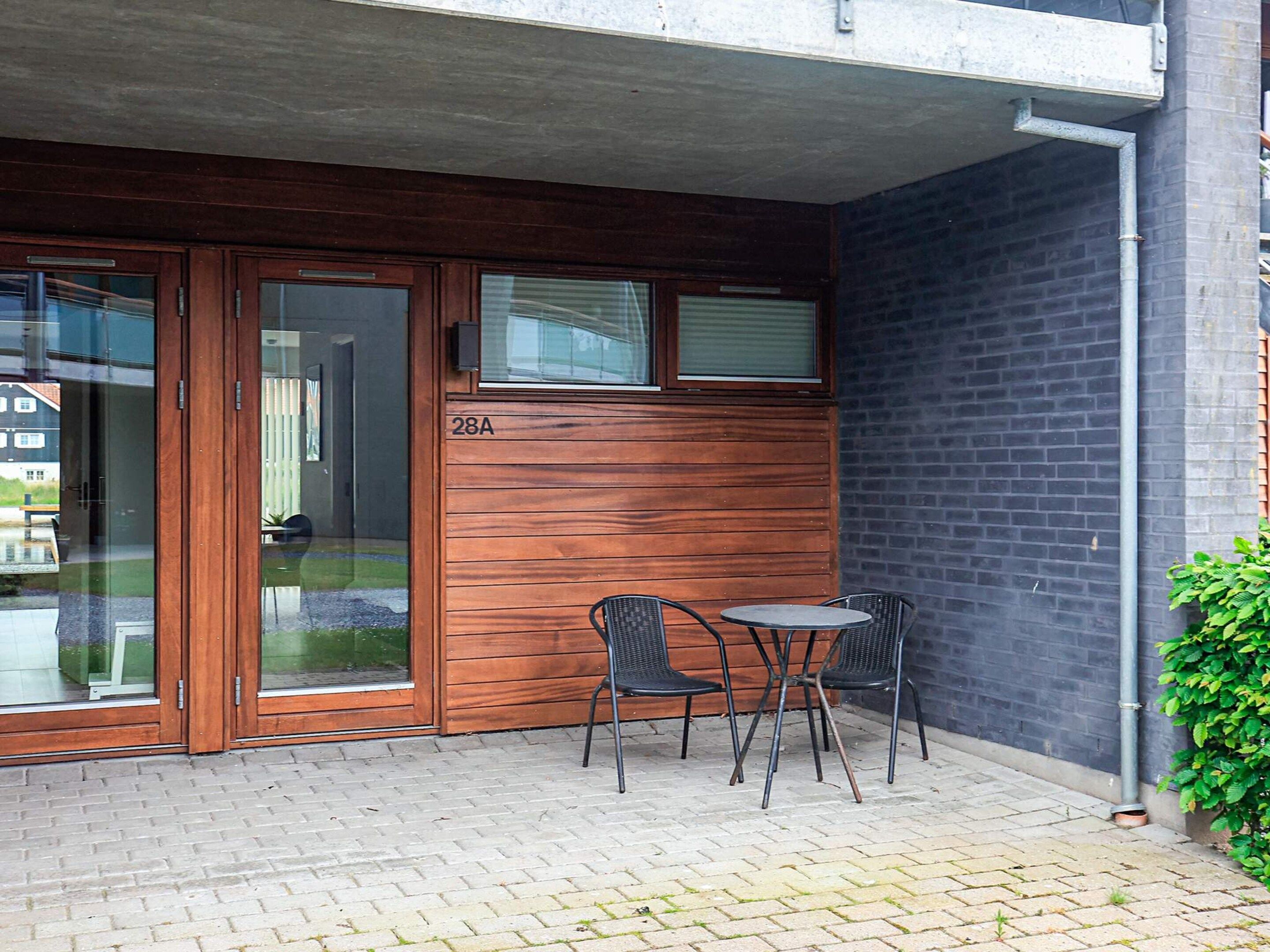 Un edificio con fachada de madera, puerta de vidrio y un pequeño espacio exterior con dos sillas y una mesa.