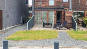 Un edificio moderno con fachadas de ladrillo y madera, un área de estar exterior cubierta con mesas y sillas, y un camino de grava.