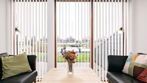 A modern living room with a wooden table, two black leather sofas, a vase with flowers, and a view of houses through large windows with blinds. - 4 Star Holiday Home in Bogense (Bogense)