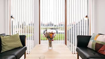 Un salon moderne avec une table en bois, deux canapés en cuir noir, un vase de fleurs et une vue sur les maisons à travers de grandes fenêtres équipées de stores.