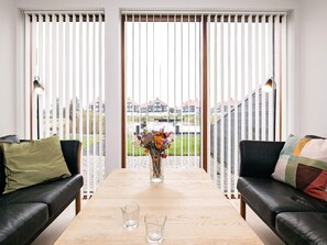 A modern living room with a wooden table, two black leather sofas, a vase with flowers, and a view of houses through large windows with blinds.